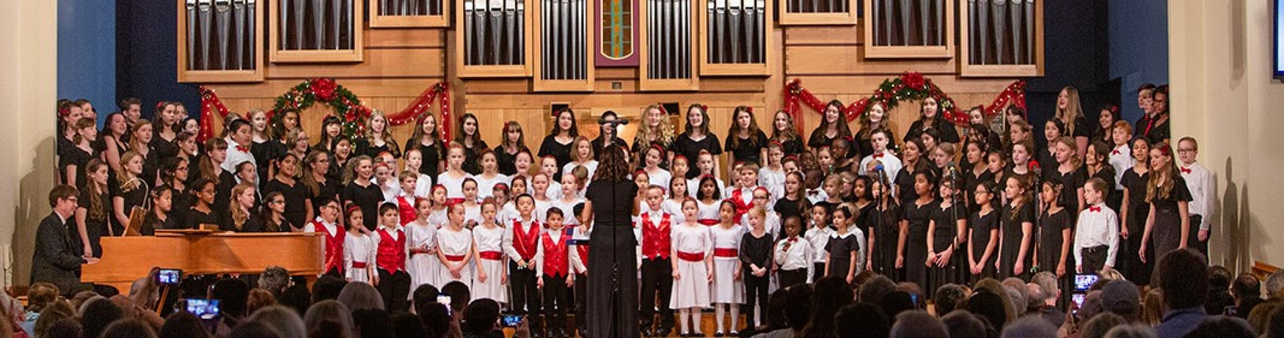 A large group of children stand on risers in front of a church organ as they sing.