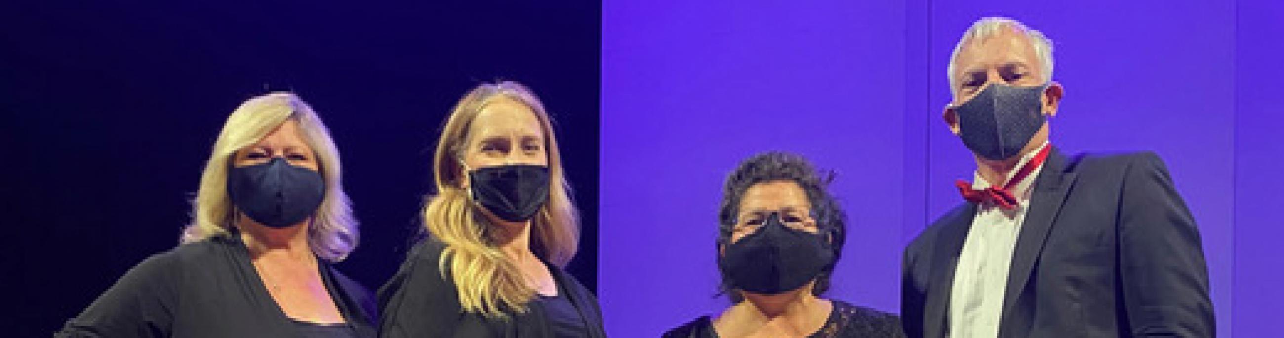 Four musicians, dressed in formal wear and masks, pose in front of a purple backdrop.