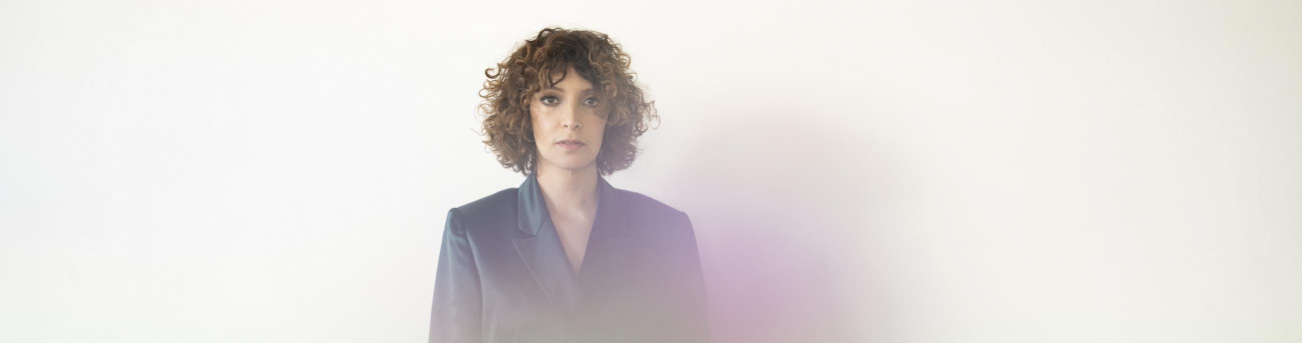 Woman with lighter skin and brown curly hair standing in the center, looking at the viewer. There is a rainbow like fog in front