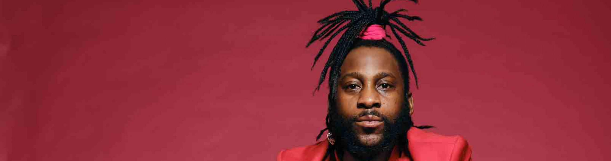 Michael Mwenso, a man with long dreadlocks in a ponytail, sits in front of a red wall in a red suit