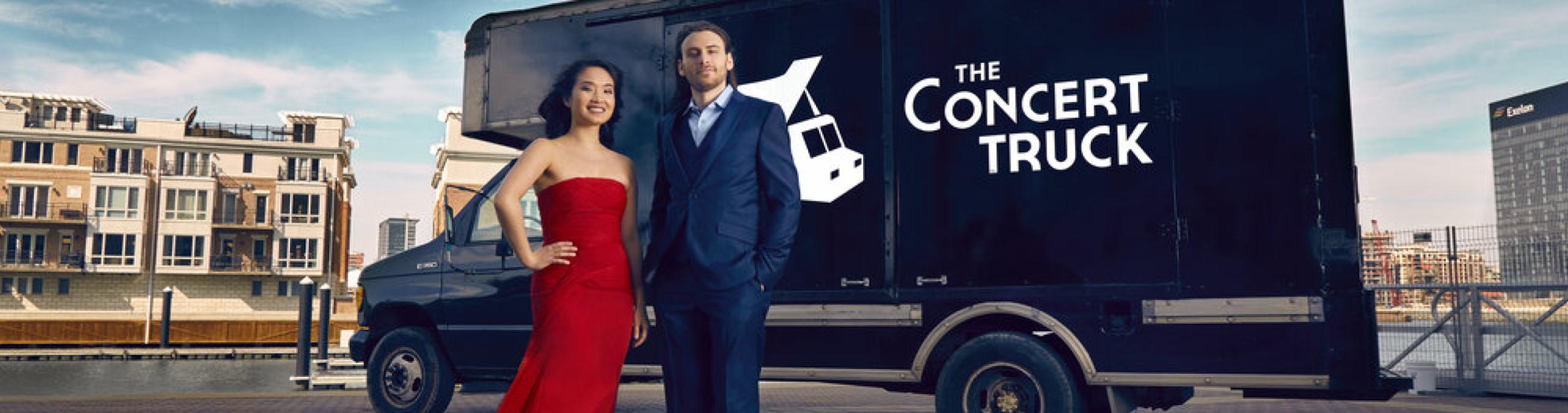 Black cargo van with the words The Concert Truck on it. A woman in a red gown and man in blue suit stand in front. 