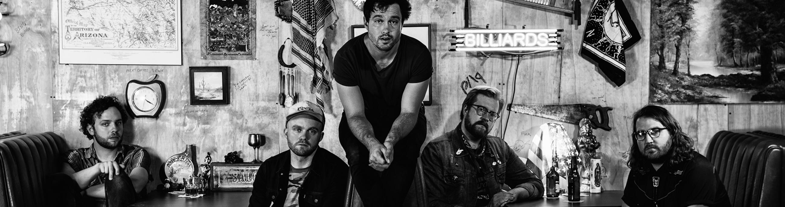 In a black-and-white photo, five male band members of Jared and The Mill sit in a booth at a diner.