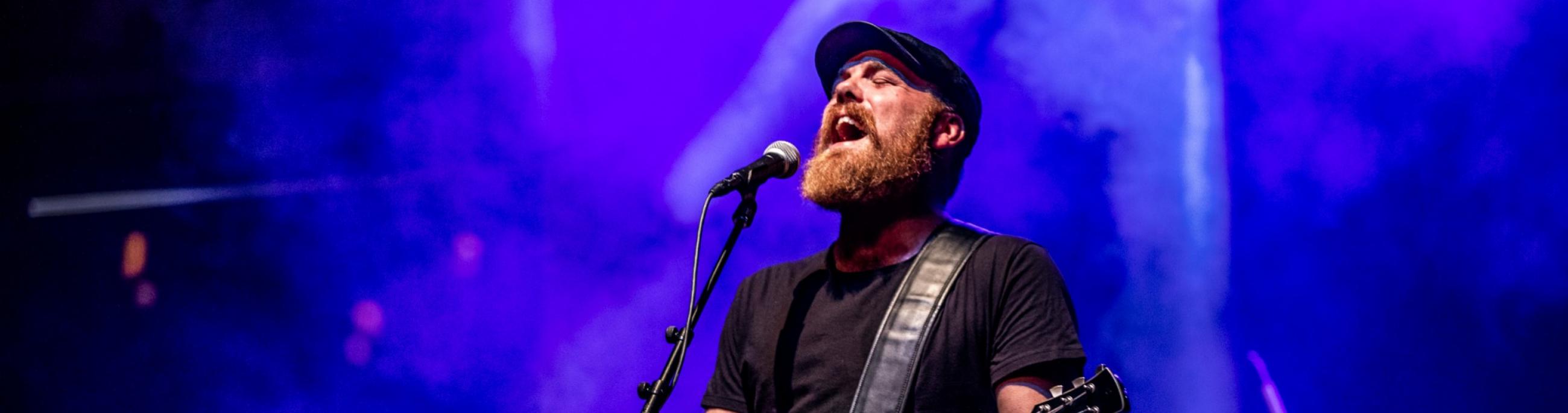 A man with a beard and black drivers cap, sings into a microphone and plays guitar