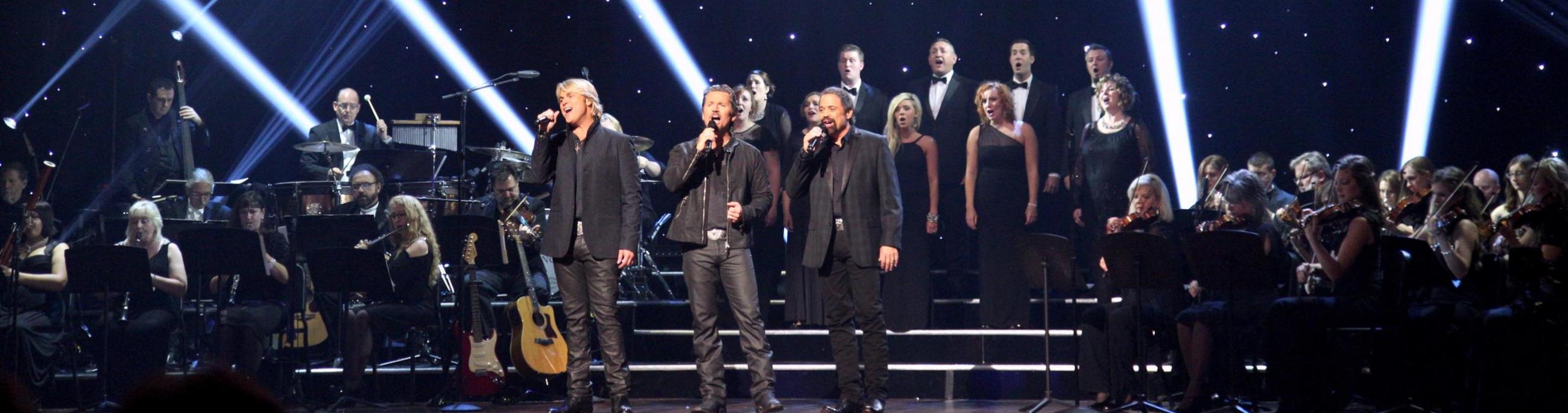 Three men with a chorus behind them standing on a dark stage singing