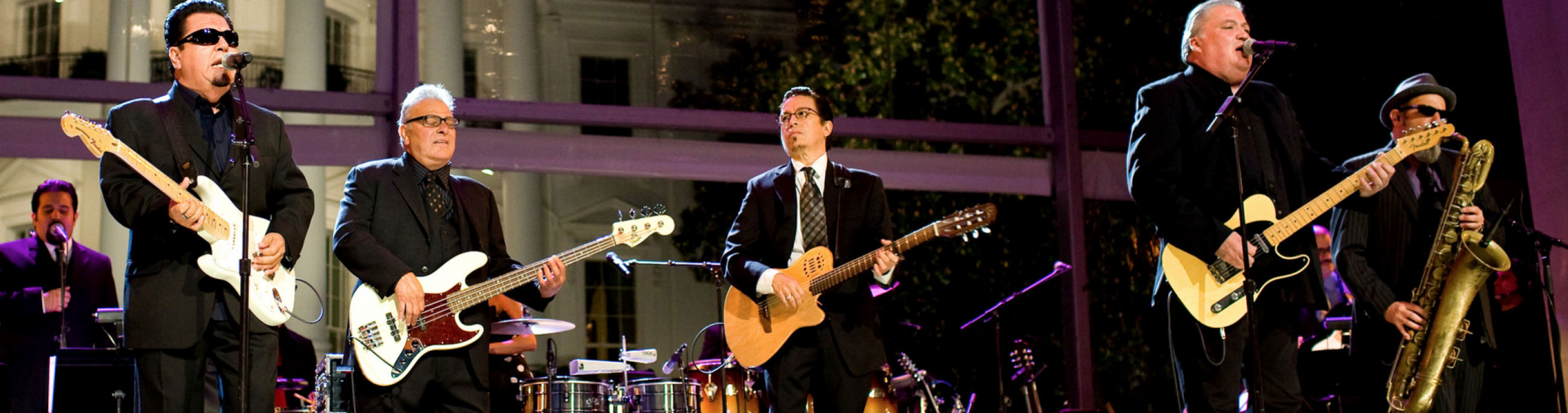 four men on stage playing instruments and singing, dressed all in black