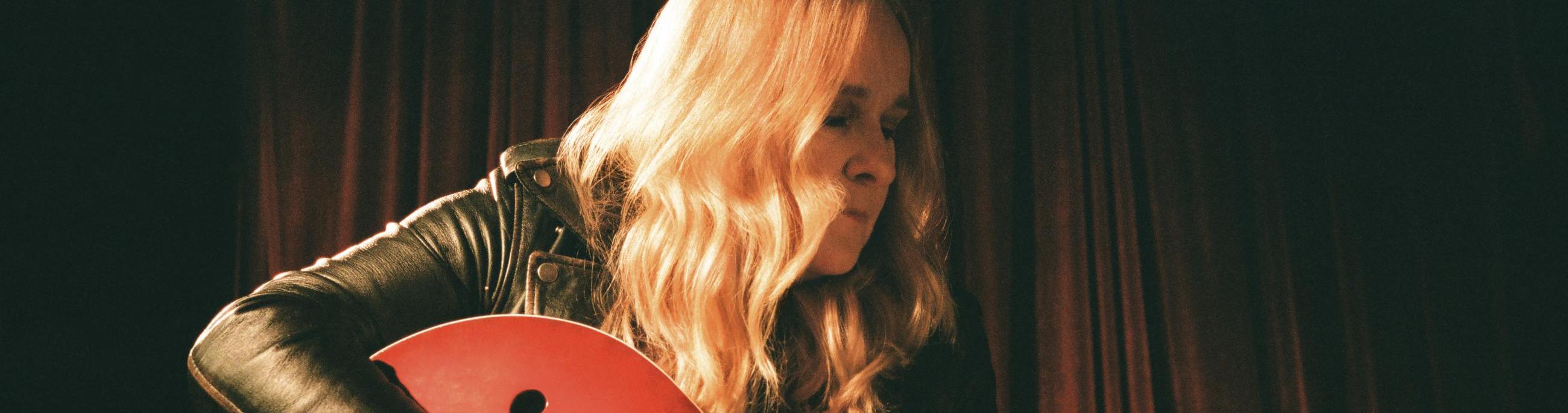 Blonde woman holding a red guitar looking to the right with a dark background