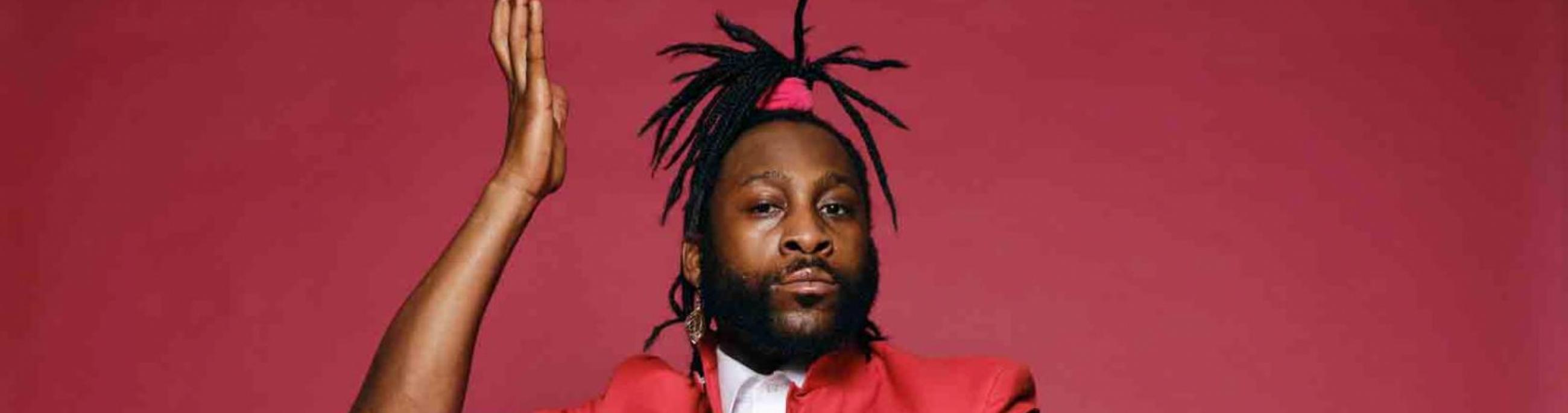 A man with dreadlocks in a ponytail wears a red suit, sitting on a chair in front of a red background. He looks at the camera.