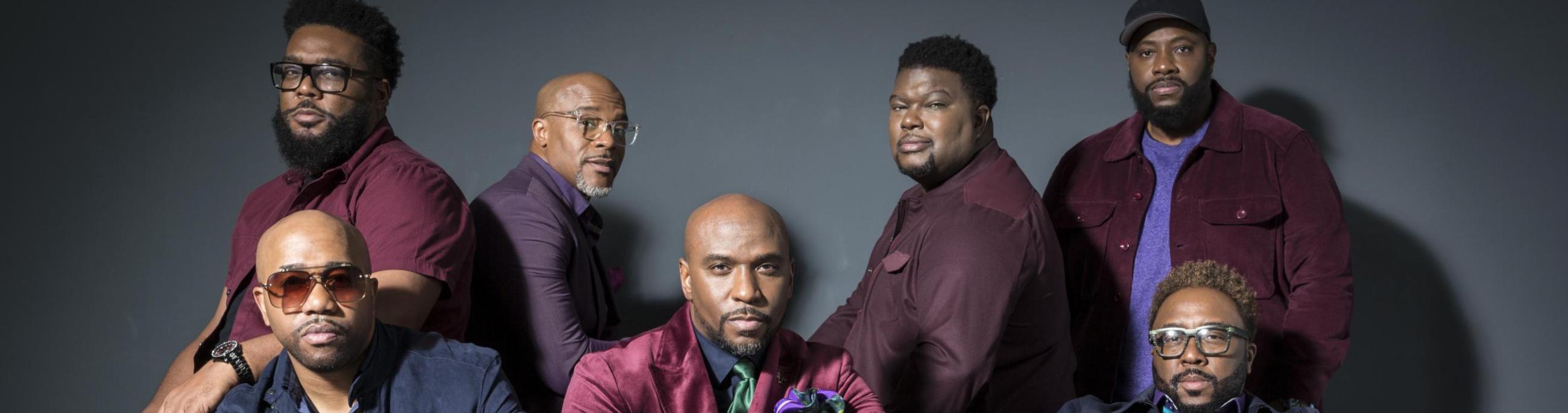 seven black men sit on a red leather couch looking at the camera