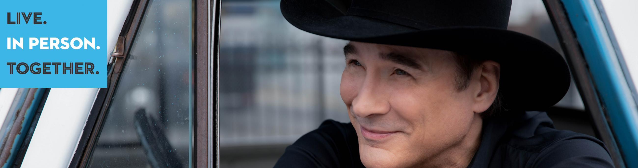 Clint Black smiles off camera as he leans out a car door. He wears a black shirt and black cowboy hat. 