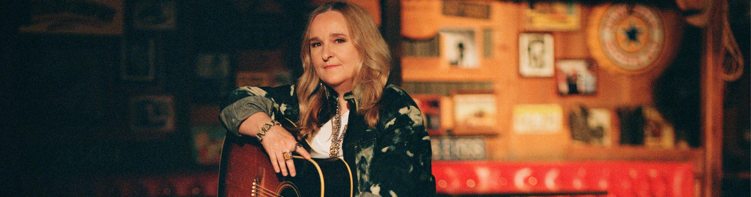 A woman with blond hair sits on a stool with her acoustic guitar on her lap, the background looks like she is in a honky tonk