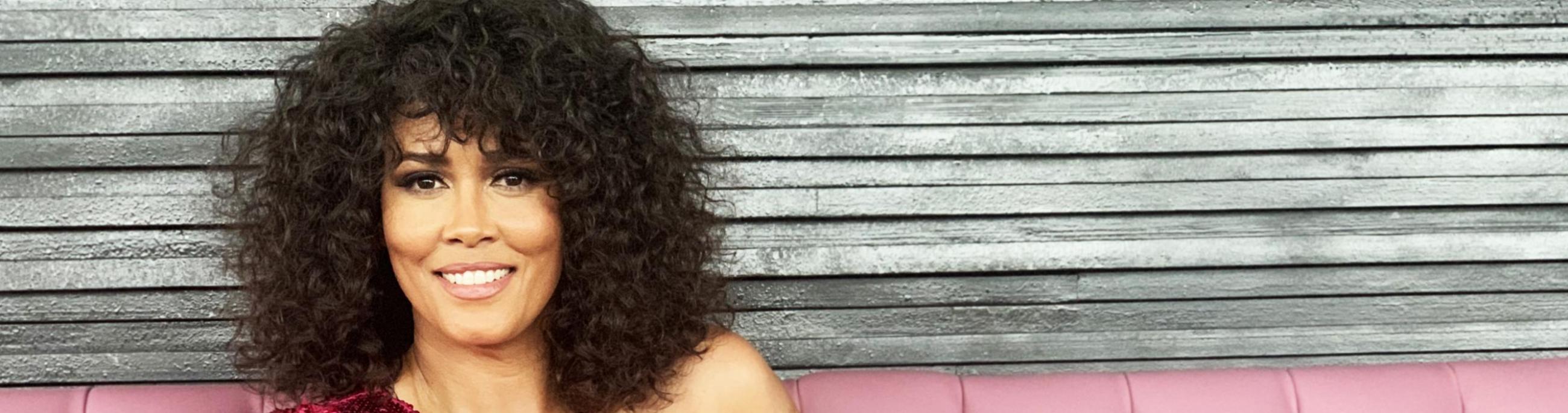 A woman with dark curly hair sites on a pink leather couch, wearing a magenta sequined top