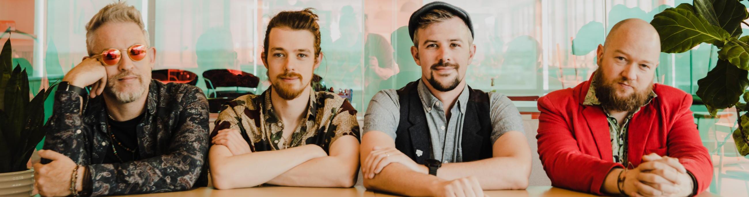 four white men sitting at a bar or table lined up and looking at the camera