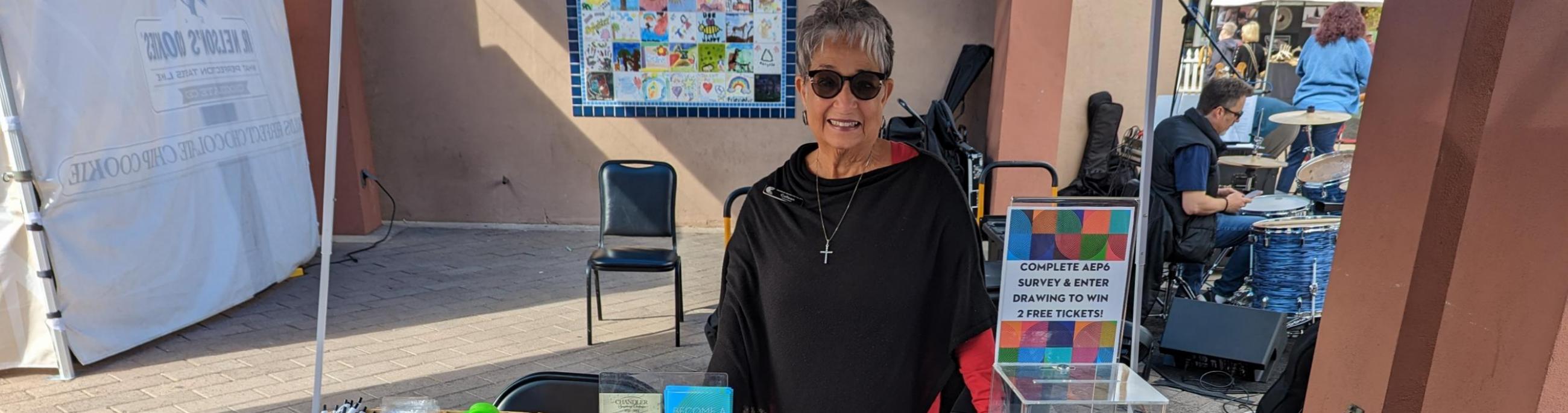 Woman in a black shirt and sunglasses standing behind a table with papers on it and a table cloth reading Chandler Center