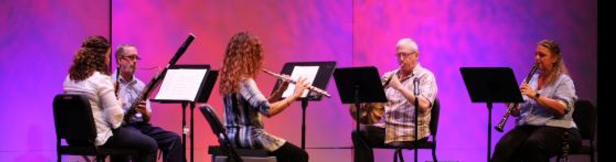 A wind quintet performs in front of a pink and purple backdrop.