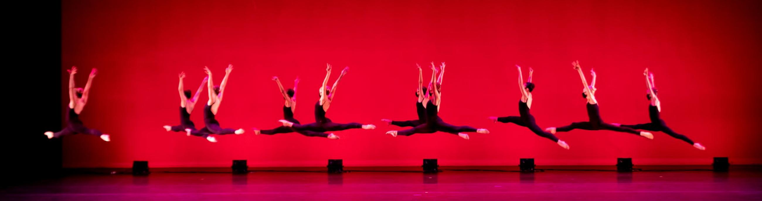 A row of ballet dancers wearing black costumes leap in front of a red background.