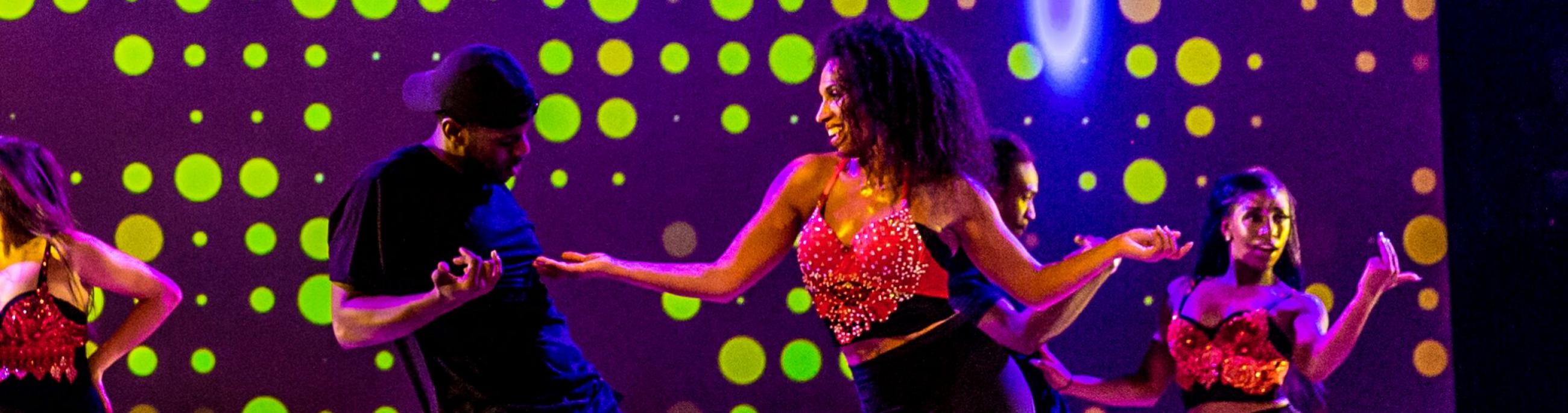 3 women and one man tap dancing, looking at each other. The lights are a purple/green and they are all wearing black and red