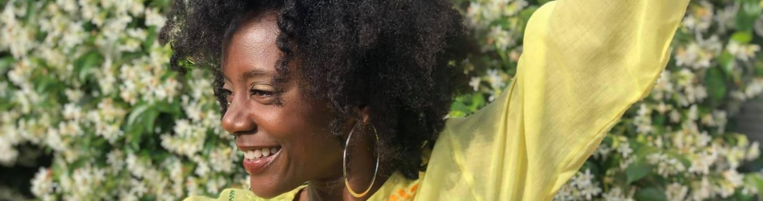 Niki J Crawford, wearing a bright yellow shirt, dances in front of a wall of white flowers.