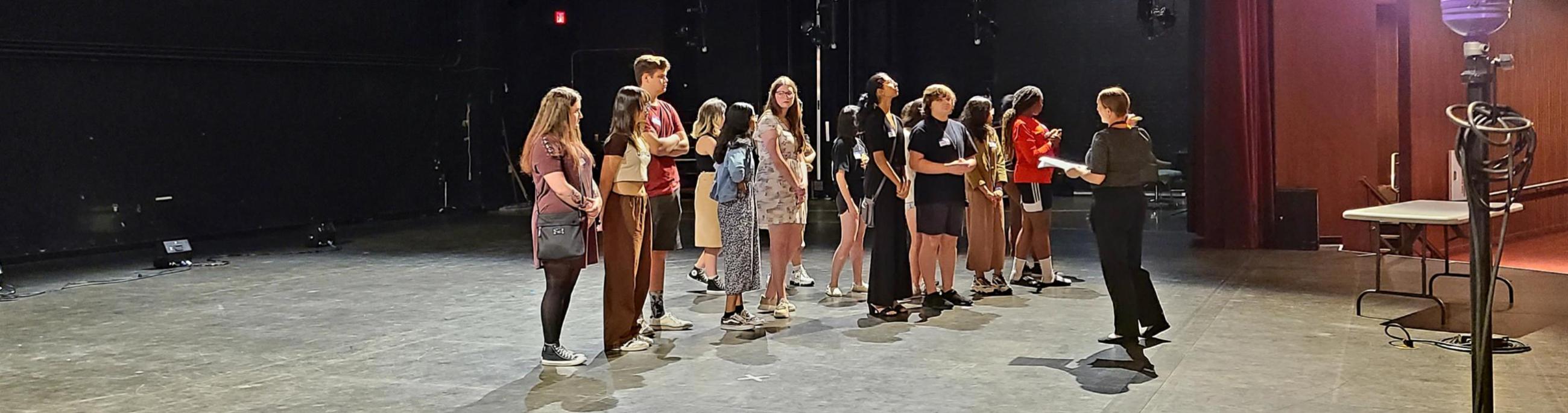 A group of students stands on a stage while a woman talks to them