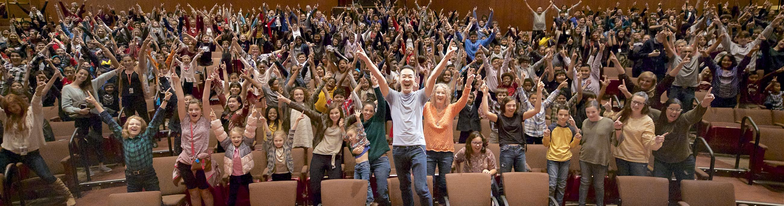 Artist Alpin Hong stands with his hands raised in joy along with a theatre full of children