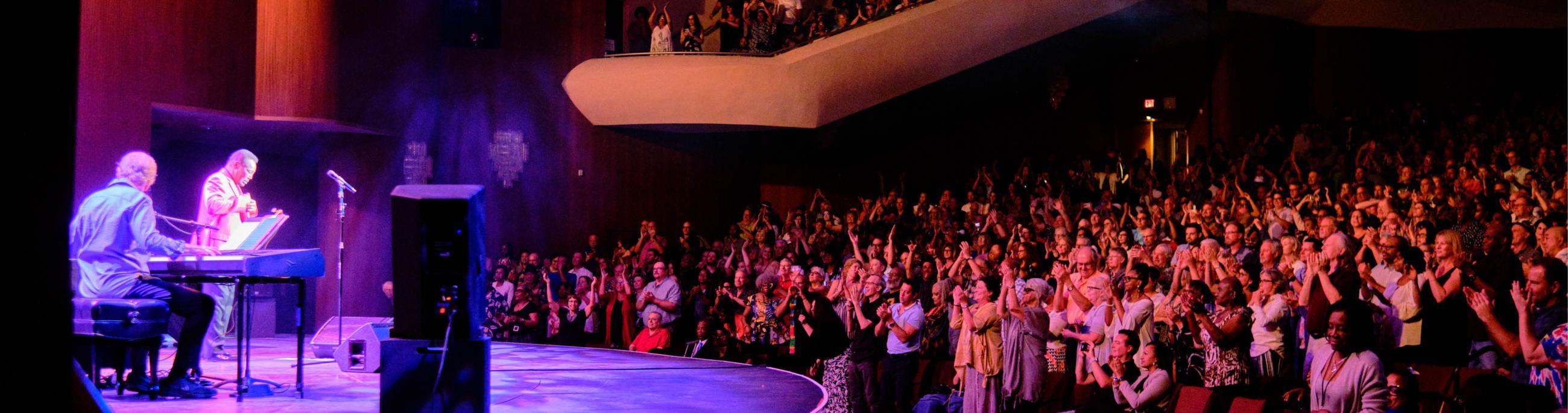 People dance and clap to George Benson who is performing on stage in a full theatre