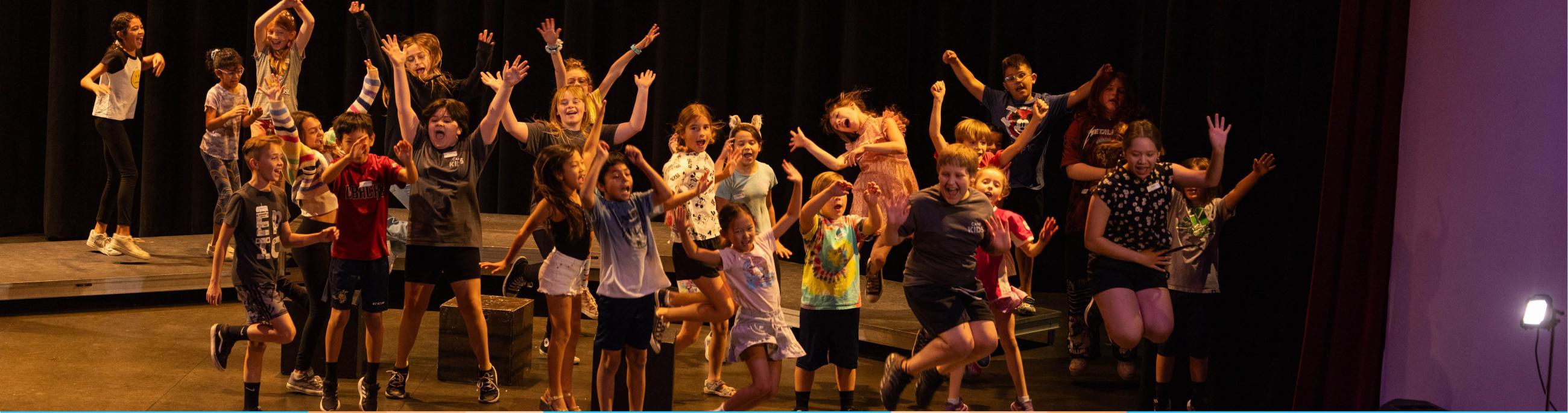 A group of youth on the theatre stage, all in a moment where they are jumping up in the air and making silly faces