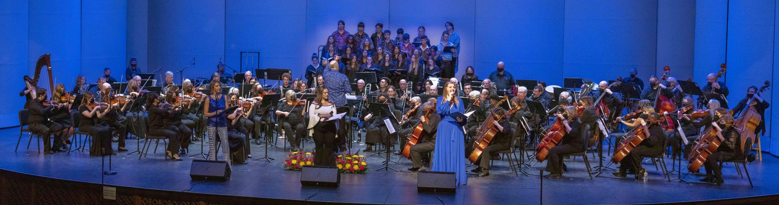 A full orchestra of musicians, conductor and two singers perform on the stage,