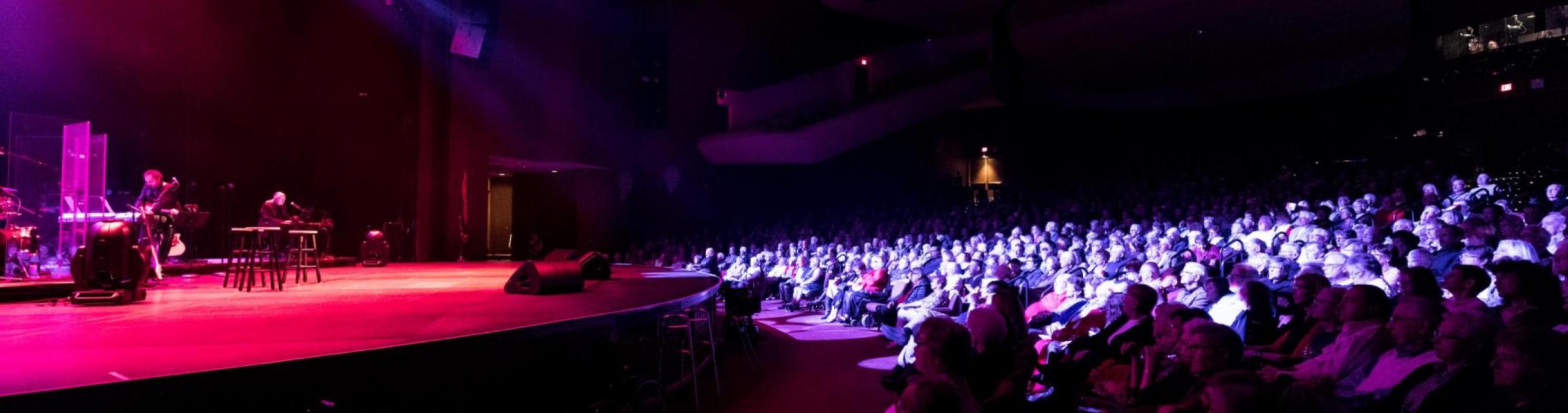 A darkened theatre, an audience watches musicians play on stage with spotlights overhead