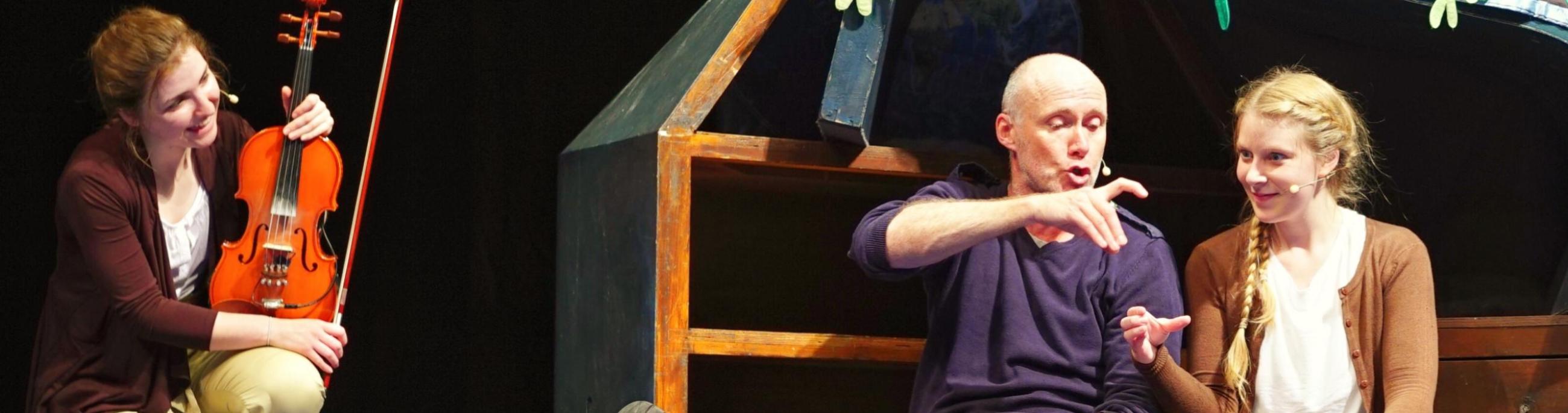  Image of young woman and older man sitting on the floor of a stage, acting out a scene as a violinist looks on