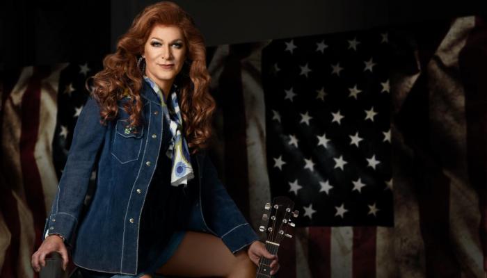 A woman with long red hair wears a jean jackets and holds the neck of an acoustic guitar