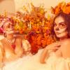 Two women pose in white dresses, orange flowers behind them and their faces painted like skeletons