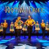 A troupe of Irish step dancers wear yellow shirts and black pants or skirts with blue lights behind them.