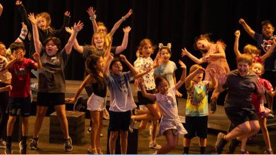 A group of youth on the theatre stage, all in a moment where they are jumping up in the air and making silly faces