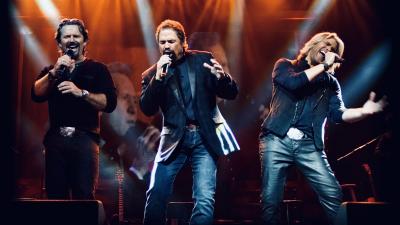 three white men singing on stage with jeans on and dark shirts with orange spotlights behind them