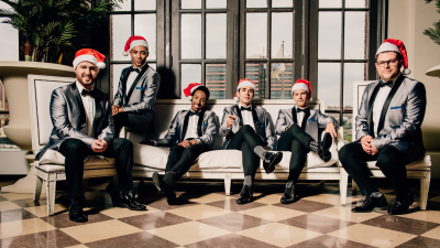 Six men dressed in black and gray suits and wearing santa hats, sitting on a couch looking at the camera