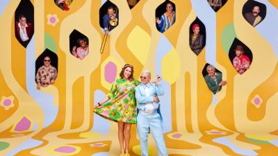 two people standing in front of a yellow background with lots of holes cut out and people sticking their heads out of the holes
