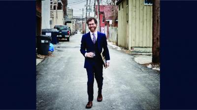 A man with glasses, a beard and brown hair wears a suit, walking in an alley.