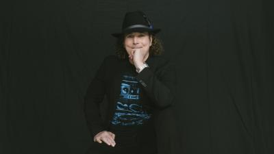 white man with curly brown hair wearing all black and a black hat in front of a black background.