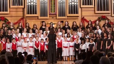 A large group of children stand on risers in front of a church organ as they sing.