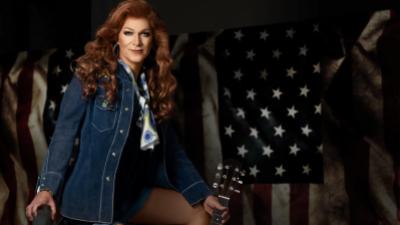 A woman with long red hair wears a jean jackets and holds the neck of an acoustic guitar