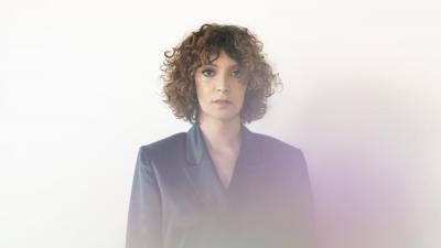 Woman with lighter skin and brown curly hair standing in the center, looking at the viewer. There is a rainbow like fog in front
