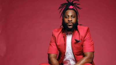 Michael Mwenso, a man with long dreadlocks in a ponytail, sits in front of a red wall in a red suit