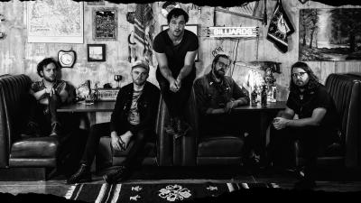In a black-and-white photo, five male band members of Jared and The Mill sit in a booth at a diner.