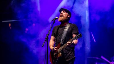 A man with a beard and black drivers cap, sings into a microphone and plays guitar