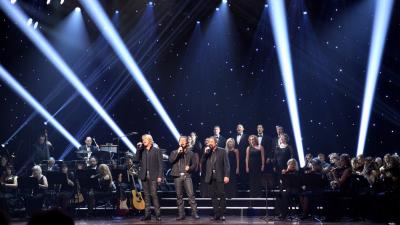 Three men with a chorus behind them standing on a dark stage singing