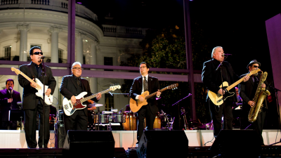 four men on stage playing instruments and singing, dressed all in black