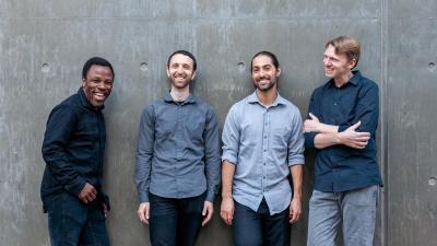 Four members of LP and The Vinyl recline against a cement wall.