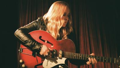 Blonde woman holding a red guitar looking to the right with a dark background