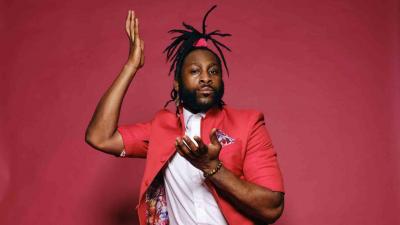 A man with dreadlocks in a ponytail wears a red suit, sitting on a chair in front of a red background. He looks at the camera.