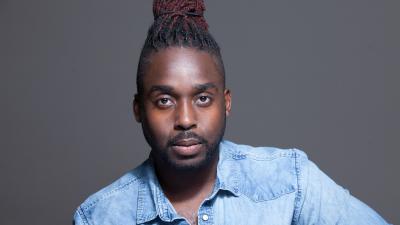 A man with a bun on top of his head looks at the camera, wearing a denim shirt