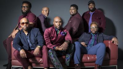 seven black men sit on a red leather couch looking at the camera